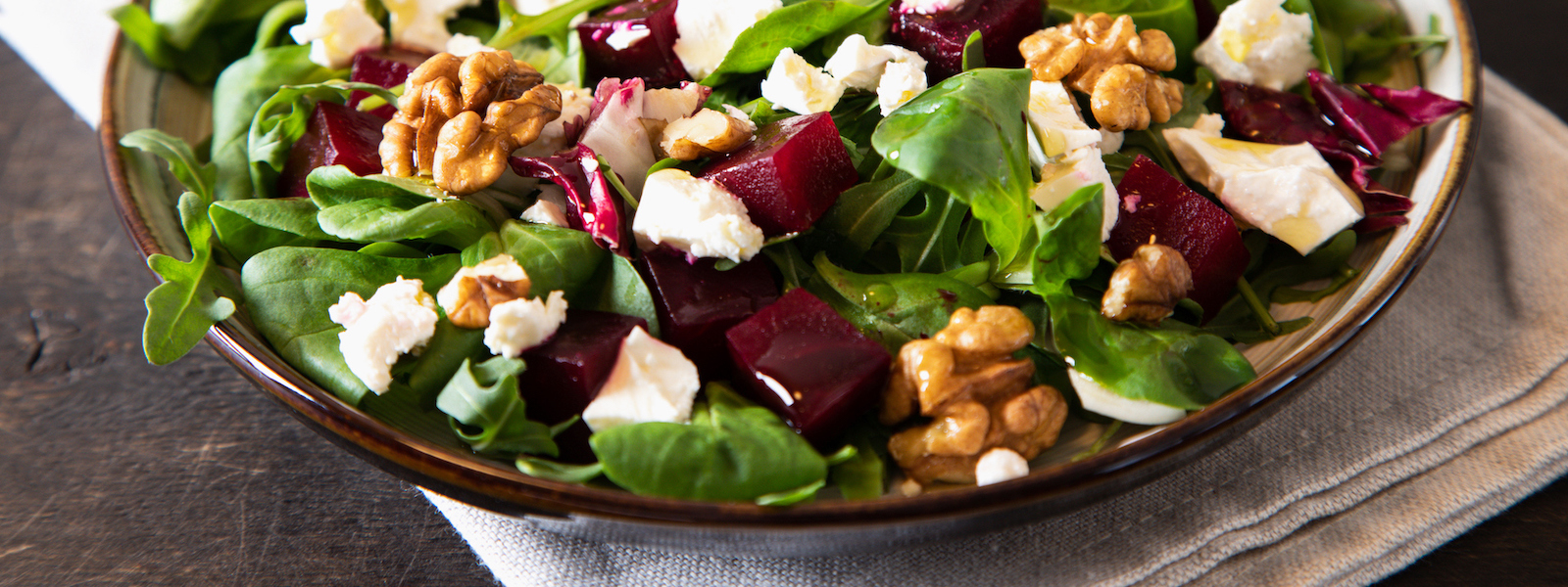 Beet or beetroot salad with fresh arugula, soft cheese and walnuts on plate, dressing and spices on dark wooden background, copy space, top view
